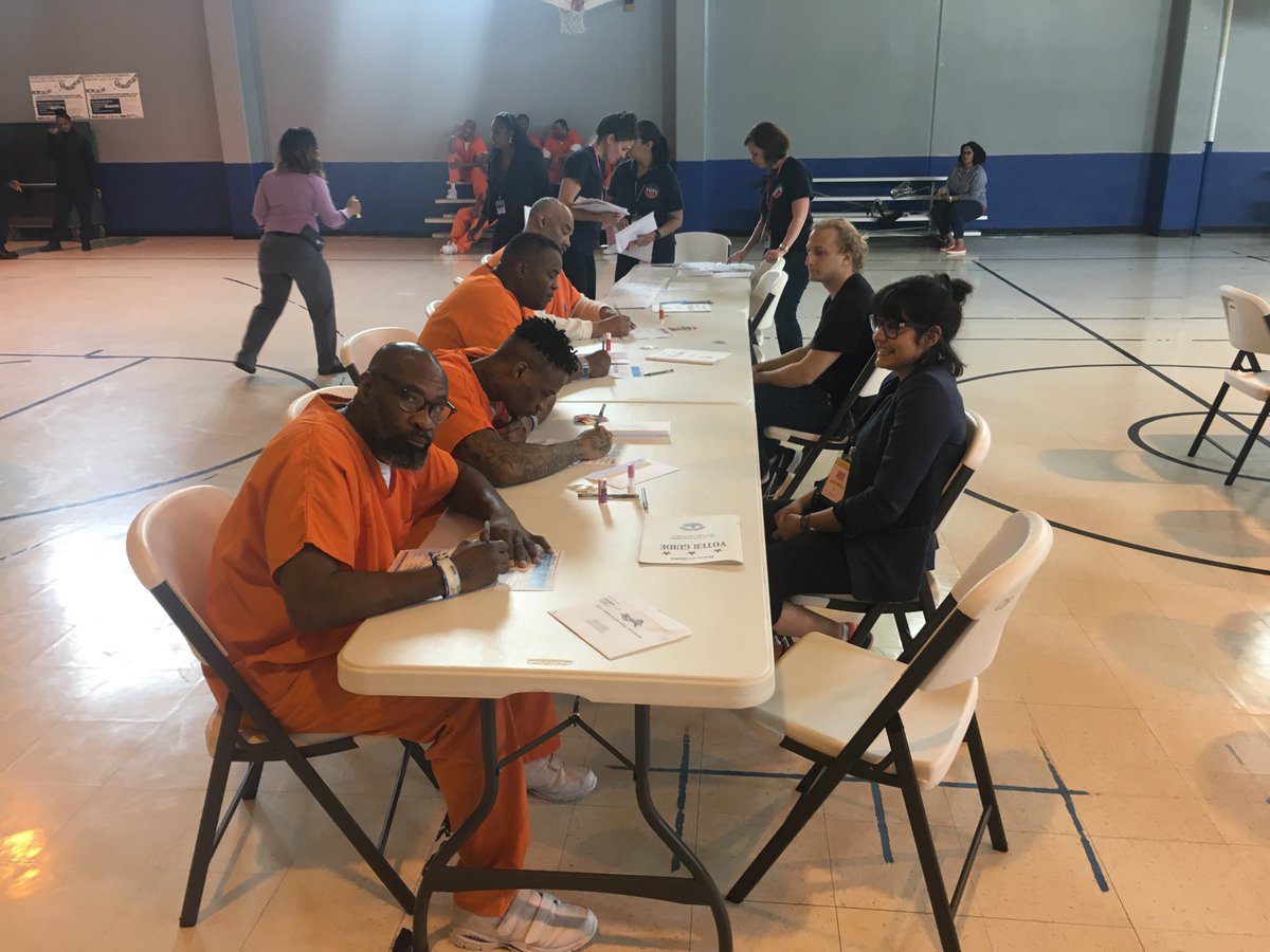 inmates in orange jumpsuits fill out ballots at a voting center table inside of a local gymnasium