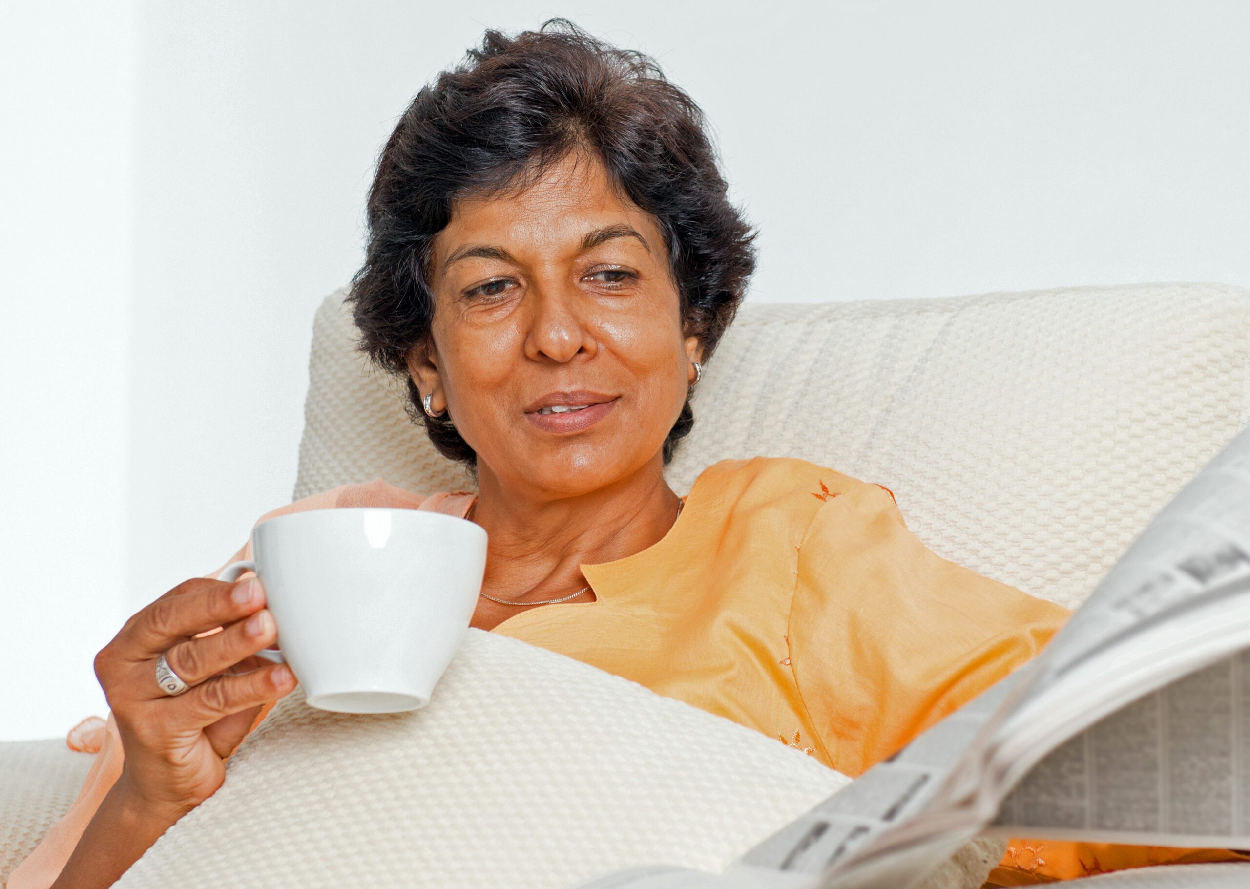 Indian woman reading newspaper and drinking coffee on a couch at home