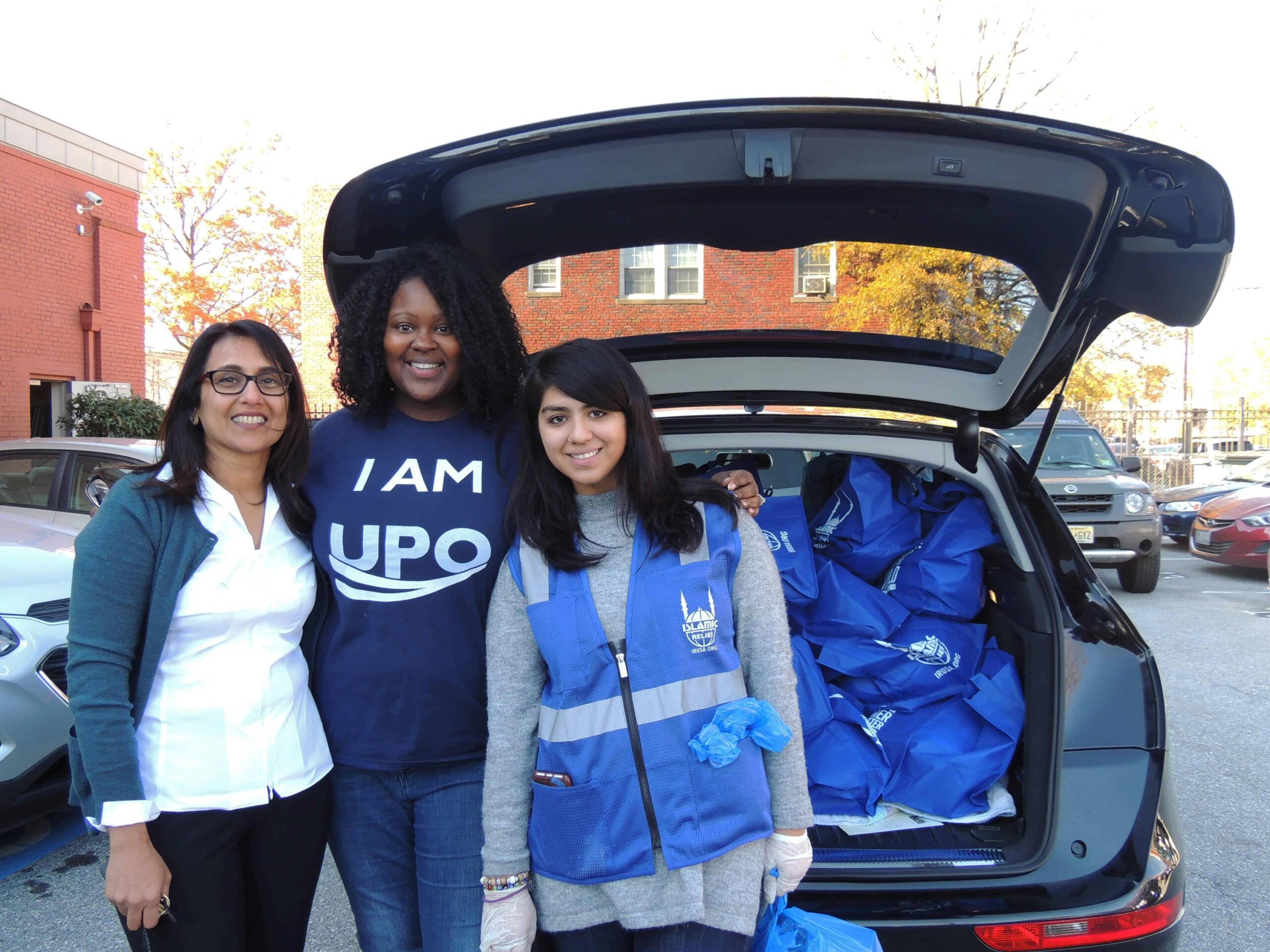 women of color smiling and posing next to car hatchback full of bags containing turkeys