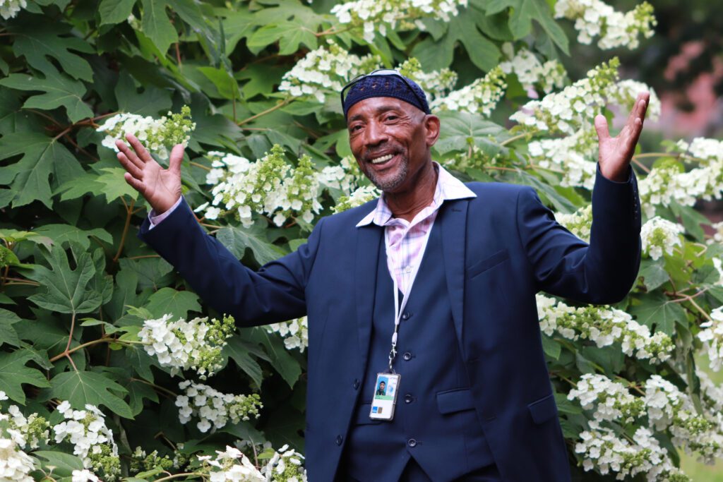 Black man smiling with hands outstretched in front of a flowering tree