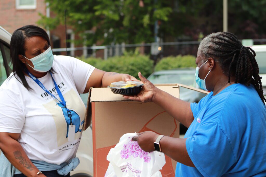 Masked UPO workers, one handing a container of food to the other